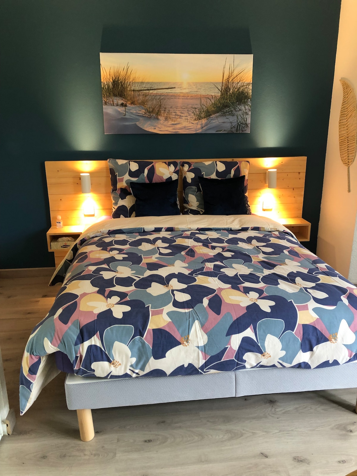 A sleeping area features a spacious bed dressed in a floral-patterned duvet cover. Two navy blue cushions are placed atop the bed's headboard, which is crafted from light wood. Soft lighting is provided by wall sconces on either side, illuminating a tranquil beach scene above the bed.