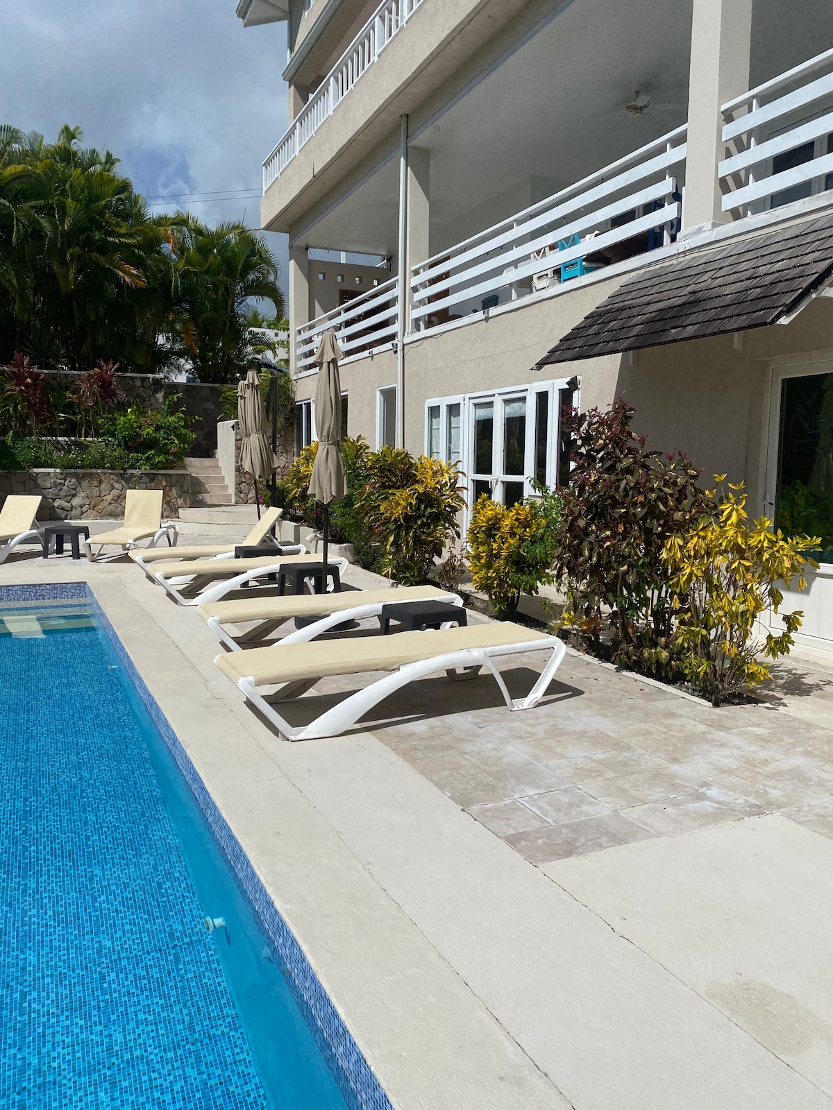 Lounge chairs are arranged beside a tranquil salt-water pool, complemented by surrounding tropical plants. A covered patio area is visible on the upper level, with umbrellas providing shade. The bright sky and greenery enhance the inviting atmosphere of the outdoor space.