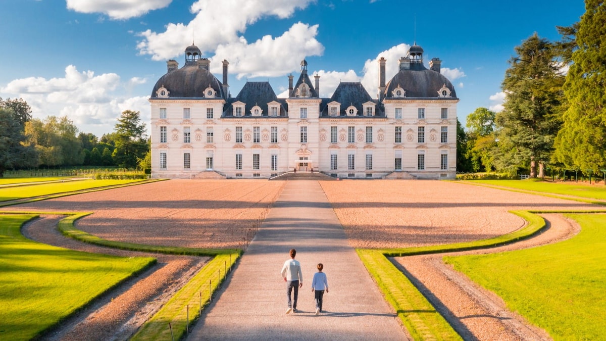 A grand chateau is viewed from the approach, showcasing elegant architecture and symmetrical gardens. The pathway leads through well-manicured lawns, flanked by circular flower beds. Two figures can be seen strolling calmly along the gravel path, surrounded by lush greenery under a bright blue sky.