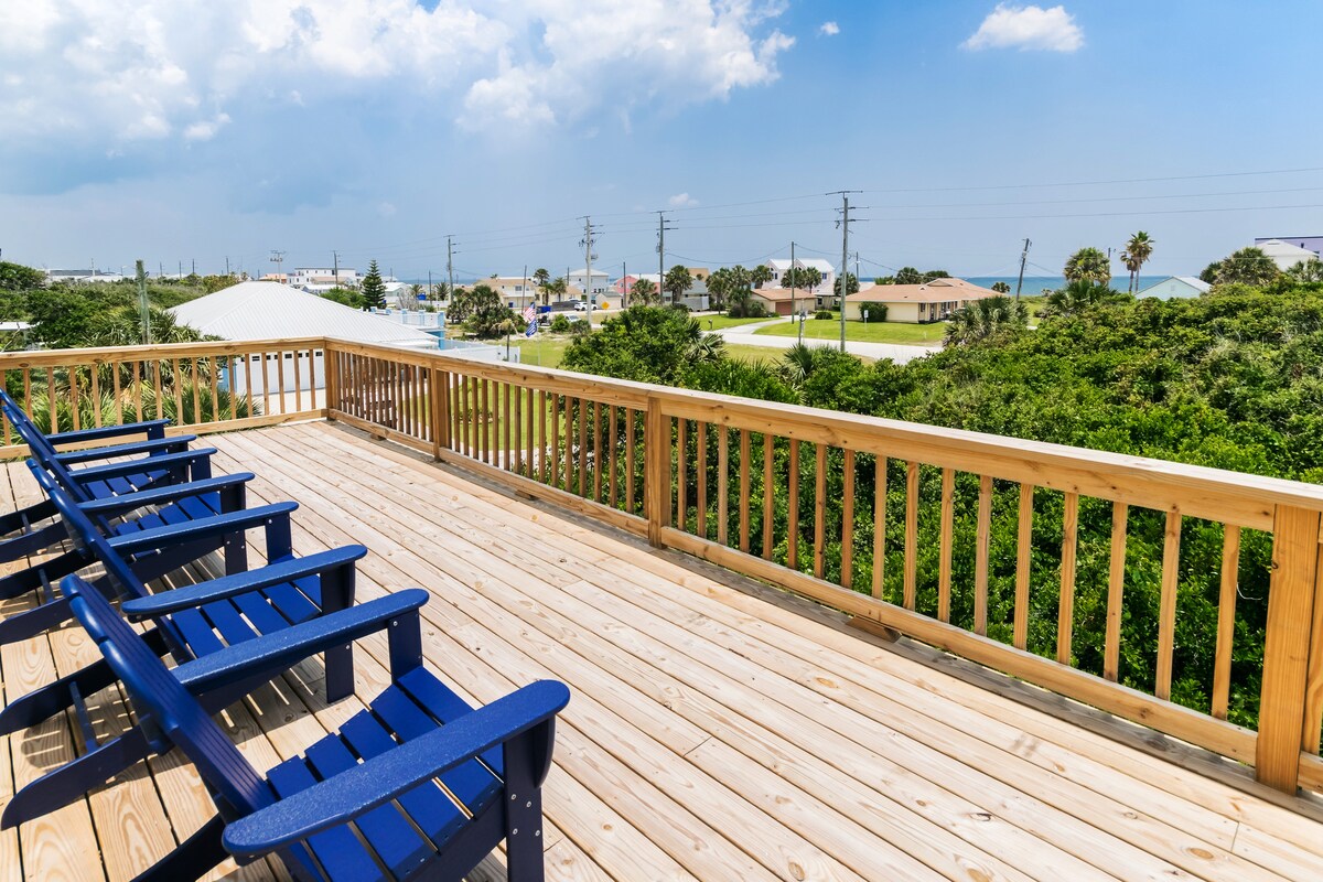 A spacious wooden deck features several blue Adirondack chairs facing a landscape of greenery and distant rooftops. The open view highlights the surrounding area, under a partly cloudy sky, providing a sense of openness and connection to the outdoors.
