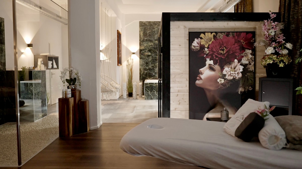 A calming wellness area is depicted, featuring a spacious treatment bed with decorative pillows. The room is accentuated by floral artwork and soft lighting, creating a serene environment. A glimpse of the private spa area is visible in the background, suggesting additional relaxation features.