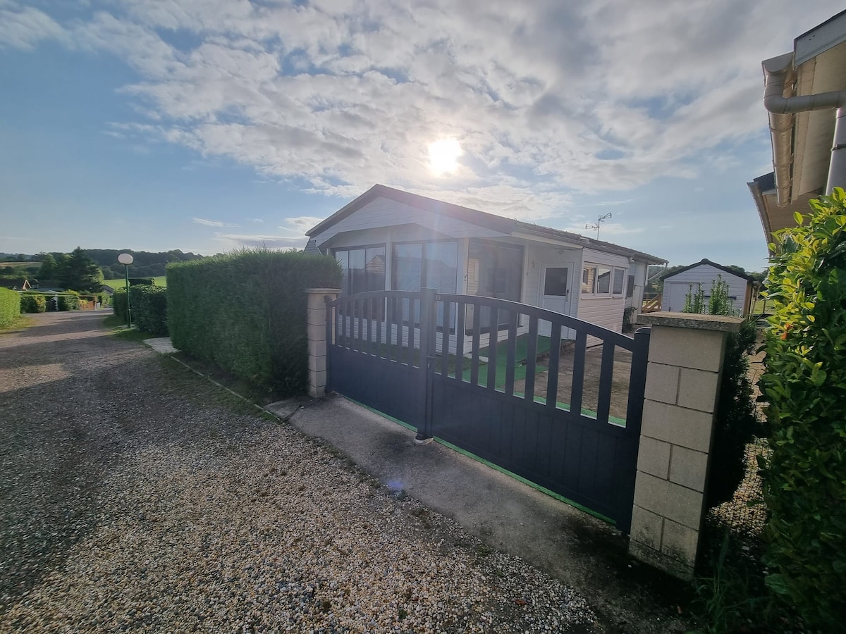 A welcoming entrance to the chalet is framed by neatly trimmed hedges and a gravel path. The gate is open, revealing a glimpse of the structure, which features large windows that allow abundant natural light. The sky is bright with scattered clouds.