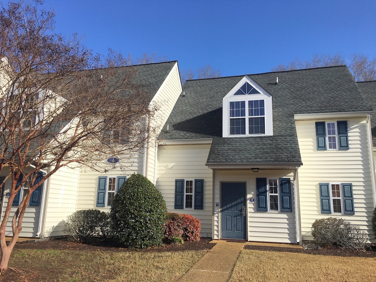 A two-story townhouse showcases pale yellow siding and a dark shingle roof. Symmetrical windows adorned with blue shutters are positioned on either side of the front door, which is framed by landscaped bushes and a well-maintained lawn.