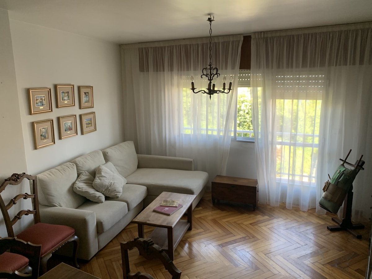 The living area features a light-colored sectional sofa adorned with soft cushions. A wooden coffee table sits in front, and a selection of framed artwork decorates the wall. Natural light filters through sheer curtains, creating a bright ambiance. Wooden flooring adds warmth to the space.