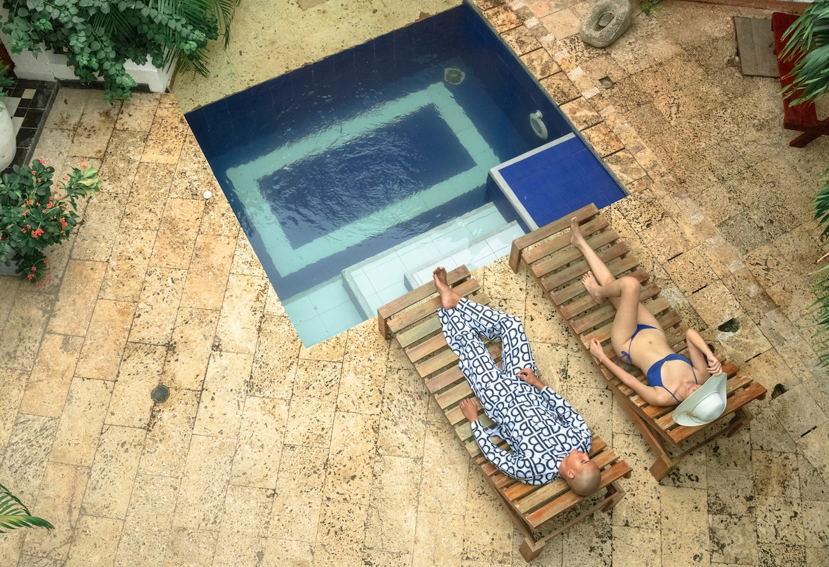 A peaceful courtyard features a rectangular swimming pool surrounded by natural stone tile. Two sun loungers are positioned on the edge, with individuals relaxing under the sun. Lush greenery adds a refreshing touch to the serene setting.