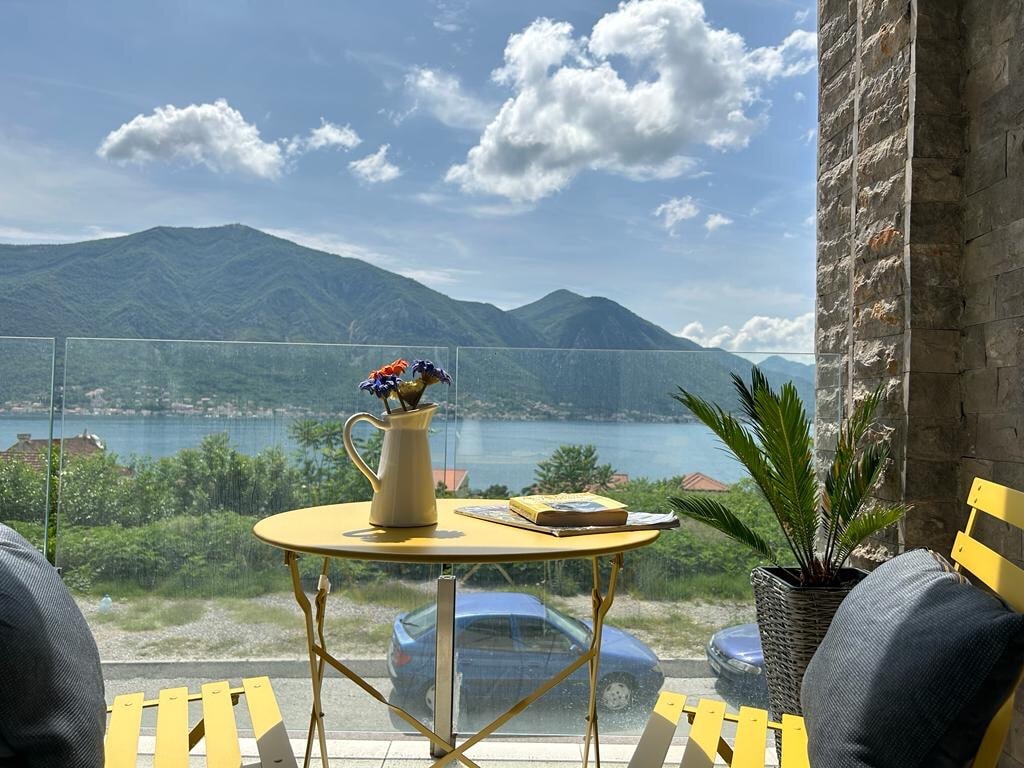 A sunlit terrace features a round yellow table adorned with a vase of flowers. Two chairs in matching yellow are positioned around the table, while a panoramic view of mountains and Boka Bay is visible in the background under a partly cloudy sky.