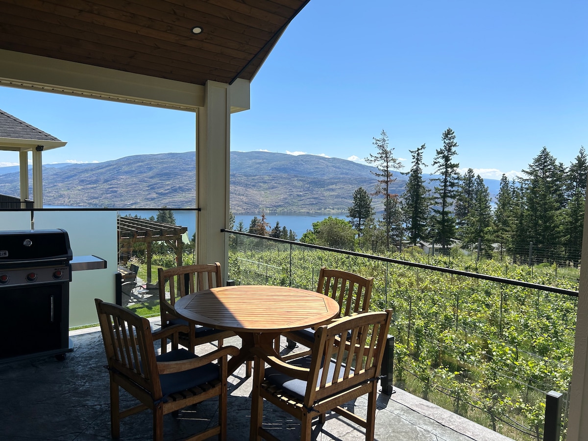An outdoor patio area features a wooden dining table surrounded by four chairs, with a barbecue grill positioned nearby. Scenic views of vineyards, mountains, and a lake are visible in the background, framed by clear blue skies and lush greenery.