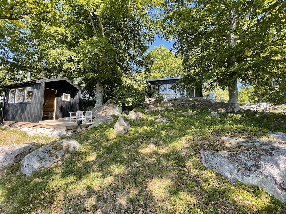 A black wooden cabin is nestled among large rocks and lush green trees, showcasing a natural hillside setting. A separate glass-walled structure is visible, complementing the rustic design. The peaceful ambiance is highlighted by the interplay of light and shade from the surrounding foliage.