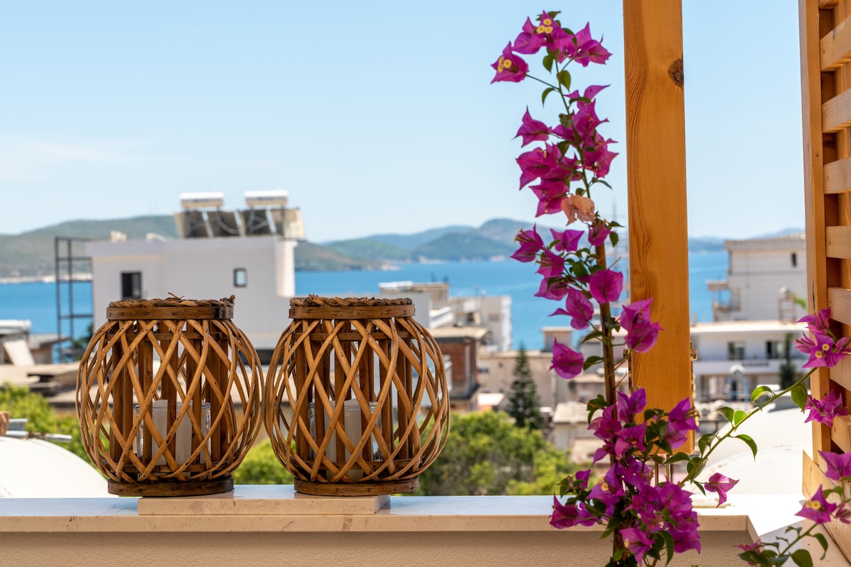 Two intricately woven bamboo lanterns are displayed on a balcony railing, accompanied by vibrant purple bougainvillea flowers. A serene view of the blue sea and distant hills is visible in the background, enhancing the tranquil outdoor setting.