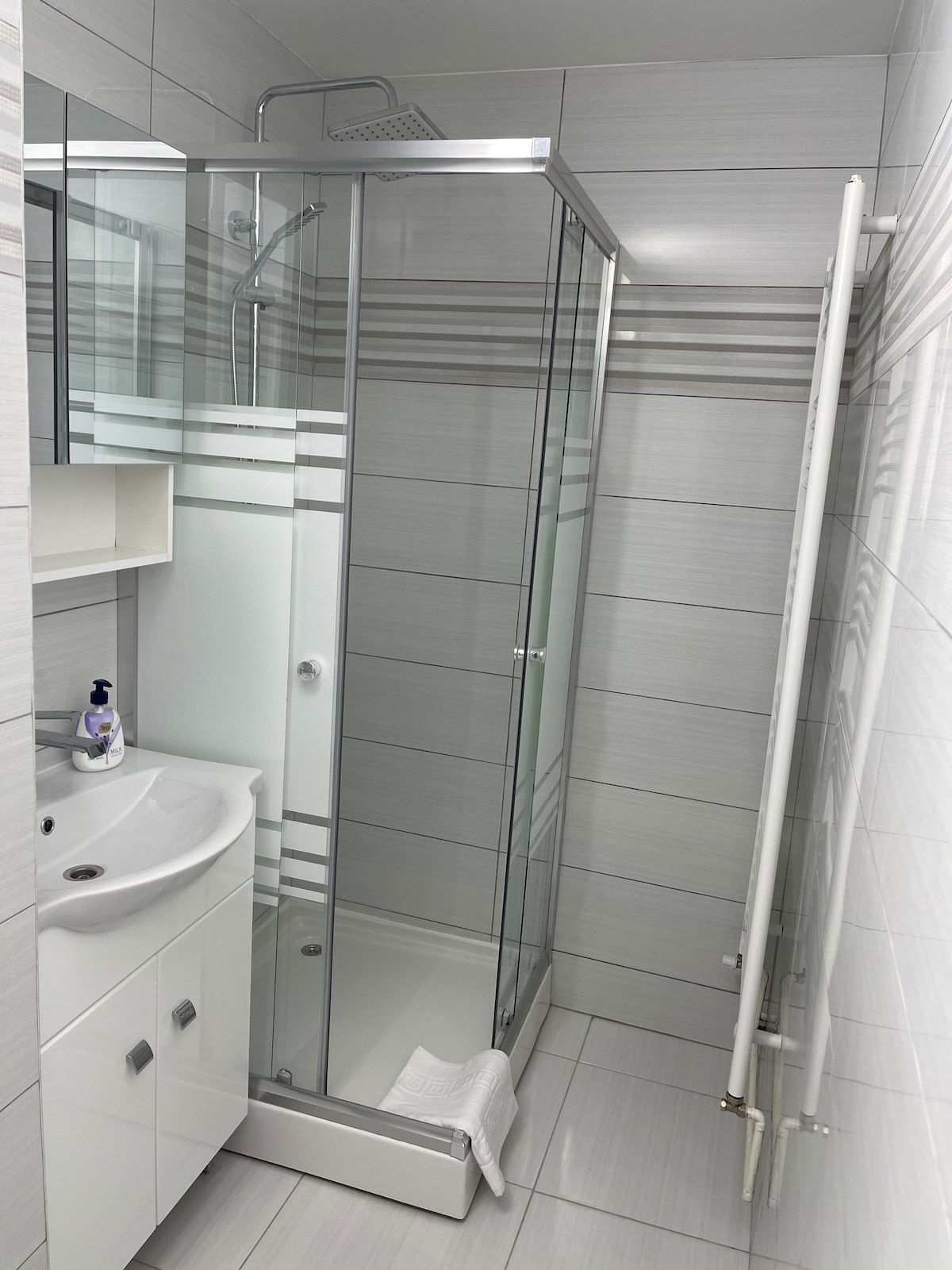A modern bathroom features a sleek walk-in shower enclosed by clear glass. A white sink is positioned beside a storage cabinet. Neutral gray tiles create a clean and fresh atmosphere, complemented by soft towels neatly arranged at the base of the shower.