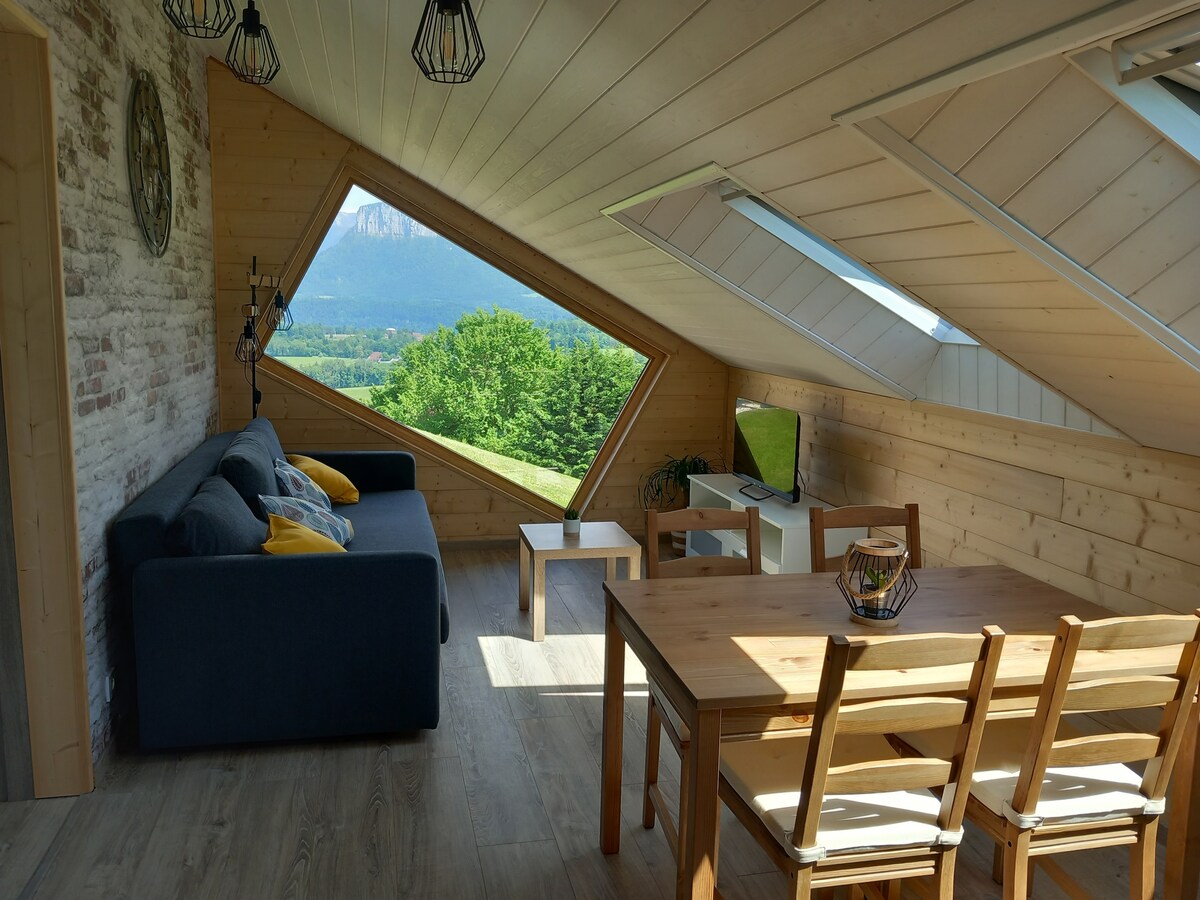 A cozy living area features a comfortable sofa, a wooden dining table, and four chairs. A slanted window offers a scenic view of the surrounding nature. Natural light fills the space, highlighting the wooden walls and flooring, enhancing the connection to the outdoors.