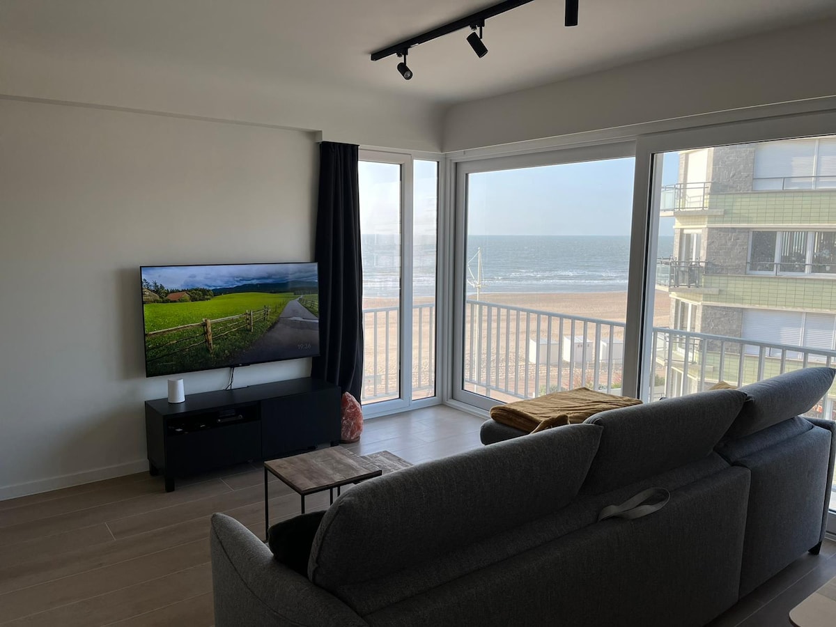 A modern living area features a gray sofa facing a flat-screen TV. Large windows offer a view of the beach, allowing natural light to brighten the space. A small coffee table sits nearby, complementing the serene coastal ambiance.