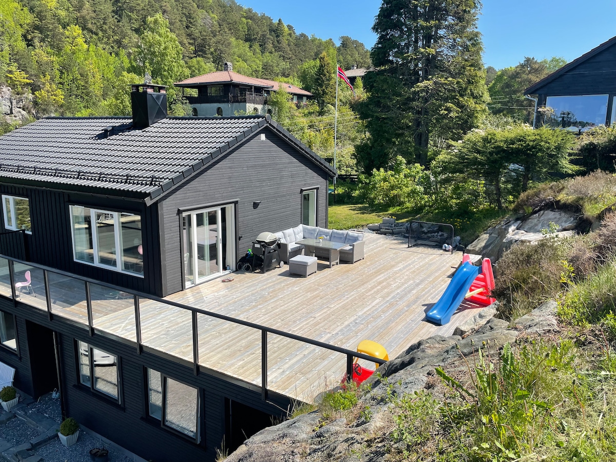 A spacious terrace is featured at the rear of the house, furnished with outdoor seating and a slide. The area is surrounded by greenery and includes a small hill, providing a serene setting. An exterior view of the house reveals large windows and well-maintained landscaping.