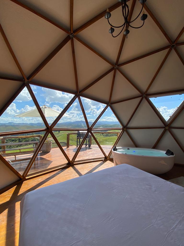 The interior of a geodesic dome features a spacious bed facing large windows that offer sweeping views of the landscape. A modern bathtub is positioned nearby, with sunlight highlighting the geometric structure of the walls and the warm wooden flooring.