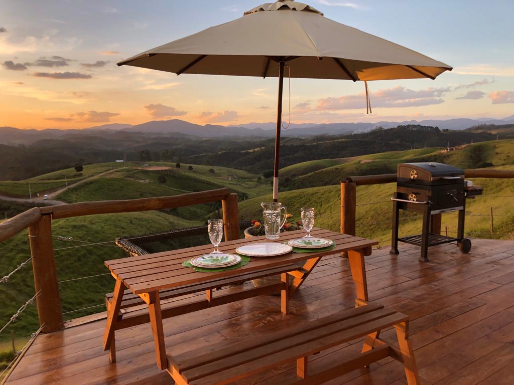 An outdoor deck features a sturdy wooden table set for dining, complete with plates and glasses, under a large umbrella. A grill is positioned nearby. The background displays rolling hills under a colorful sunset, creating a serene setting for meals.