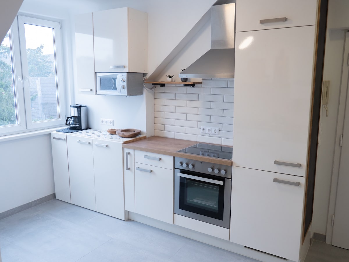 A modern kitchen is presented, featuring sleek white cabinetry and stainless steel appliances. A combination oven and stovetop are complemented by a microwave and coffee maker on the countertop. Natural light enters through a large window, illuminating the tiled backsplash.