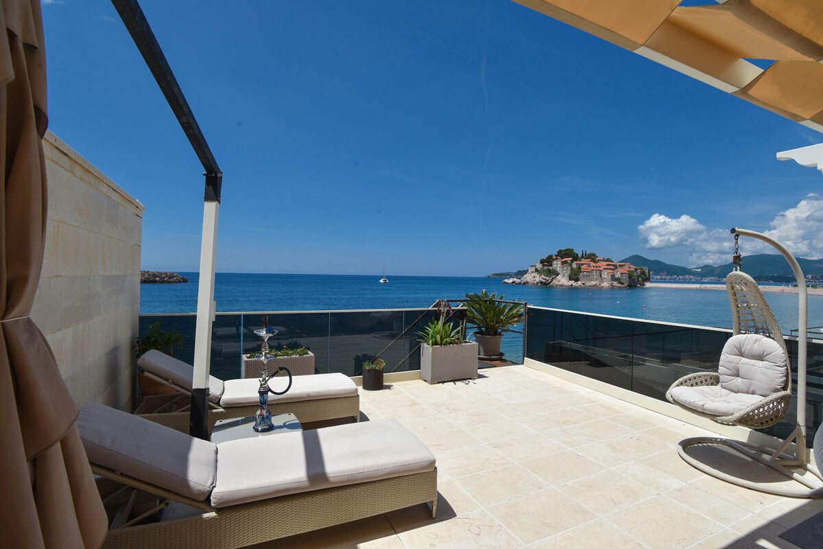 A spacious terrace overlooks the sea, featuring sun loungers and greenery. The tranquil waters reflect the blue sky, while Sveti Stefan island is visible in the distance. An elegant seating area invites relaxation, providing an ideal space to enjoy the remarkable views of the coastline.