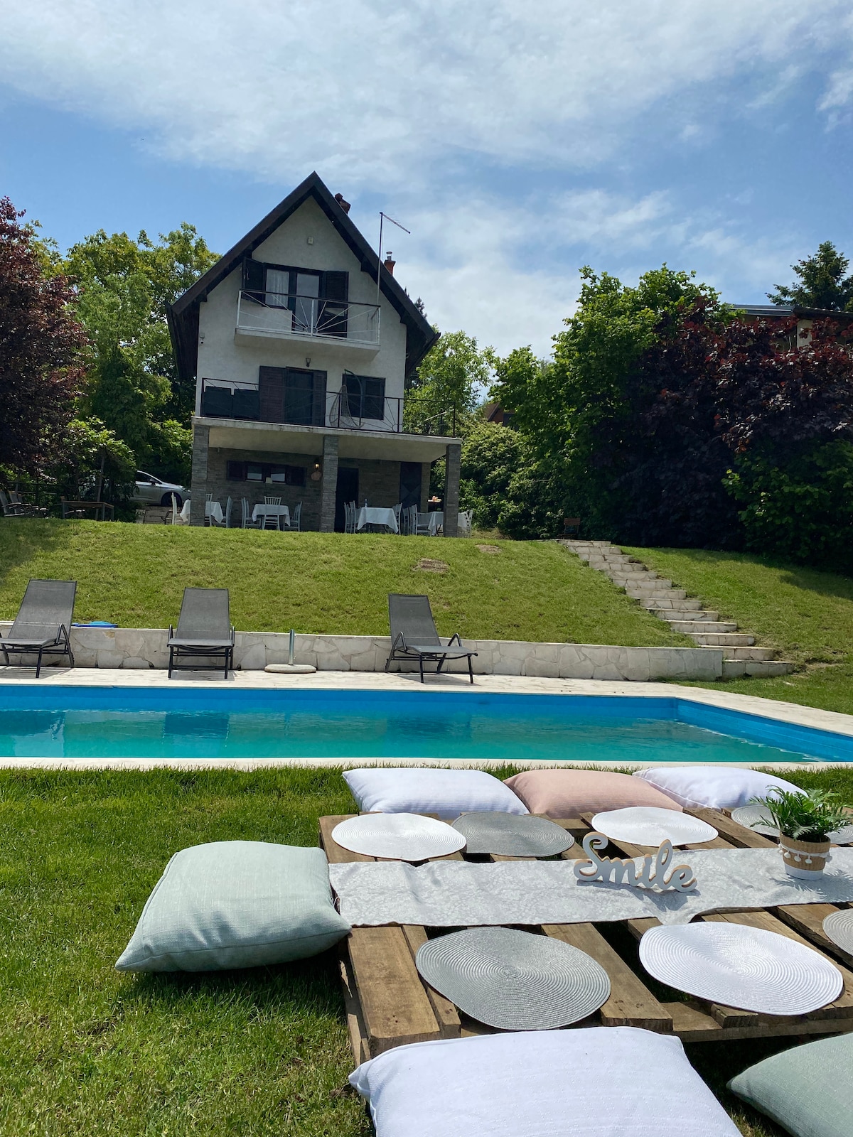 A spacious outdoor area features a inviting pool surrounded by green lawn. Lounge chairs are arranged near the pool. The two-story house is visible in the background, complemented by a patio area with multiple seating options and steps leading to the yard.