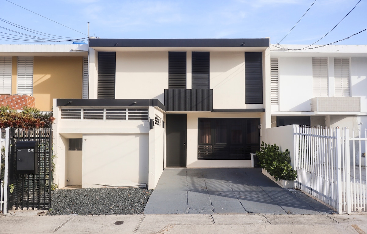 A modern, two-story house is showcased in the image, featuring a clean design with horizontal lines. The façade includes large windows and a dark entryway that contrast with the lighter exterior. A driveway leads to the garage, accompanied by pebble landscaping on one side.