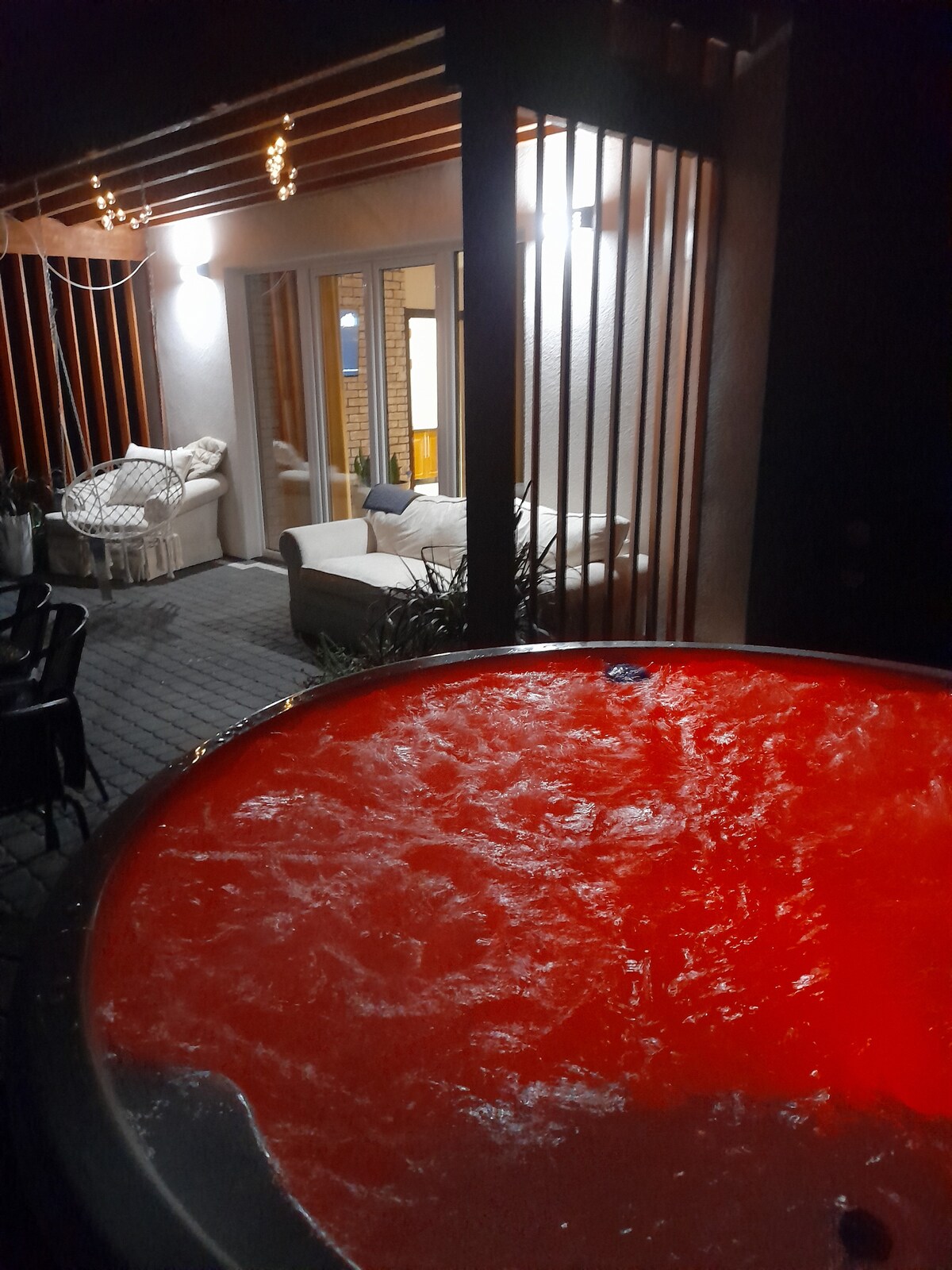 A hot tub with illuminated red water is positioned in the foreground, surrounded by a stone patio. In the background, a cozy seating area is visible, featuring a white couch and decorative pillows, partially shadowed by the wooden slats of the structure.