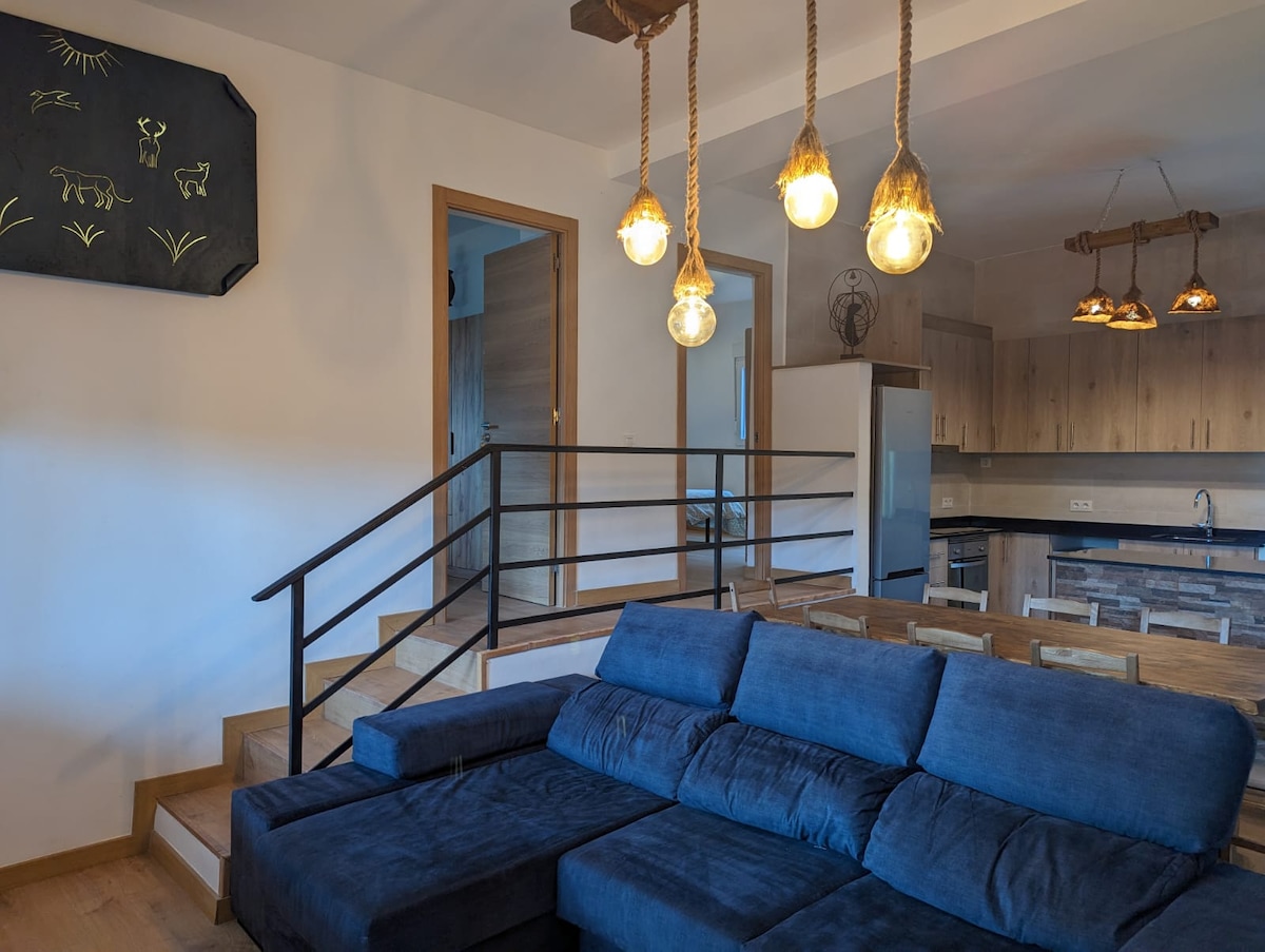 A spacious living area is highlighted by a dark blue sectional sofa, inviting relaxation. Warm pendant lights hang above, illuminating the room. Stairs lead to additional levels, while an open-plan kitchen is visible in the background, featuring wooden cabinetry and modern appliances.