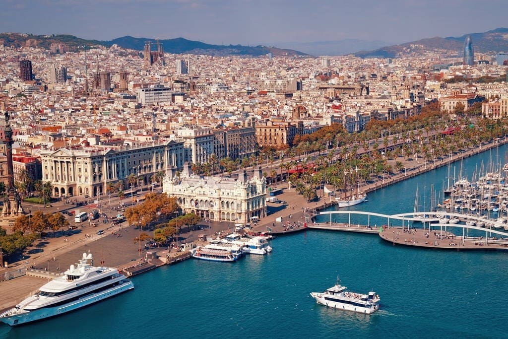 An aerial view captures the vibrant cityscape of Barcelona, featuring a bustling marina with various yachts and sailboats. The coastline is lined with palm trees, while historical buildings and modern architecture coexist along the waterfront, creating a picturesque urban landscape.