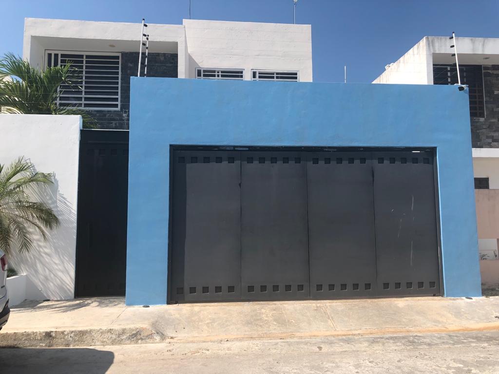 A modern building features a vibrant blue exterior wall with a large black garage door that dominates the front. Lush green foliage is visible to the left, providing a contrast to the clean lines of the structure. The sky above is clear and bright.