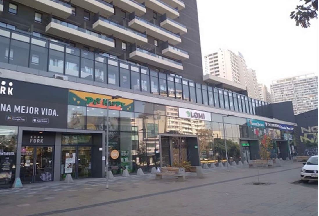 A modern building with a glass facade showcases various storefronts on the ground level, including a minimarket and restaurants. The surrounding area features paved walkways and small trees, providing a pleasant urban environment. Tall buildings loom in the background, typical of a cityscape.