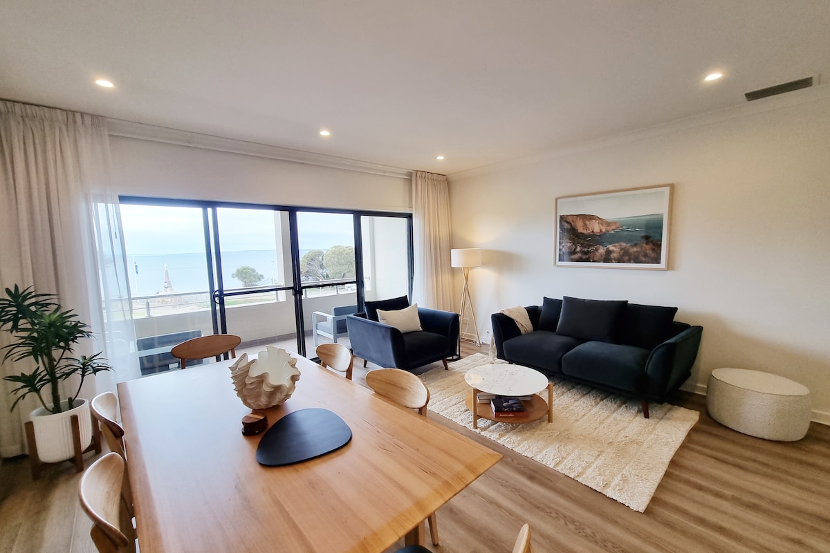 The open-plan living area features a dining table surrounded by light-colored chairs and a cozy seating arrangement with a dark sofa and armchairs. Large windows offer views of the ocean, creating a bright and airy atmosphere.