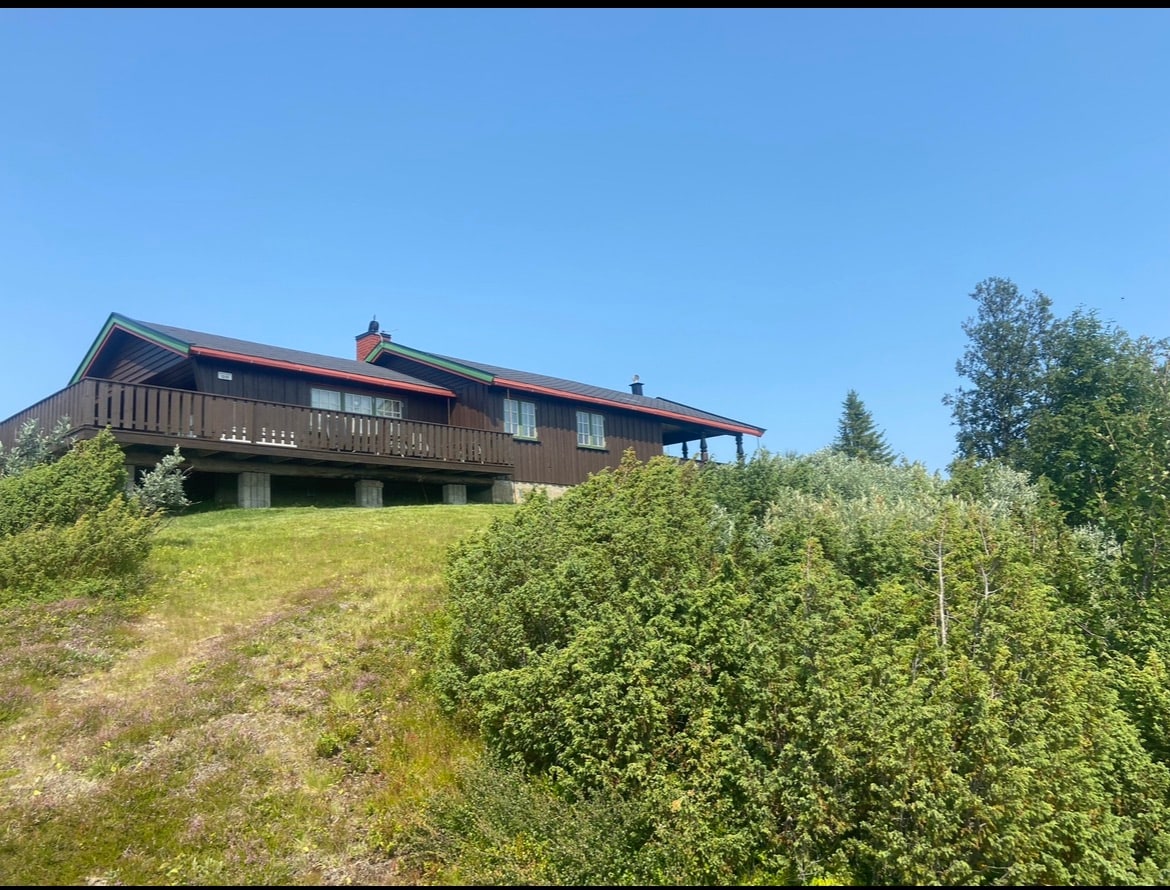 A spacious cabin is positioned on a green hillside, showcasing an expansive deck that offers views of the surrounding landscape. The building features a gabled roof and multiple windows, allowing natural light to fill the interior. Lush vegetation frames the cabin, enhancing its secluded feel.