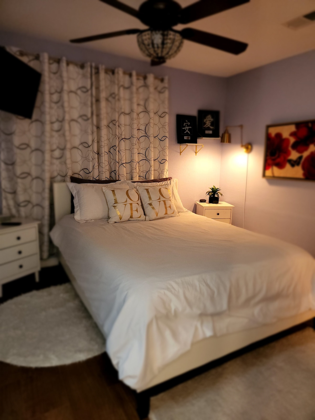 A serene bedroom is featured, showcasing a spacious bed adorned with decorative pillows. The walls are painted in a soft lavender, complemented by elegant curtains. A small nightstand with a lamp and a potted plant adds a touch of greenery, while wall art enhances the visual appeal.