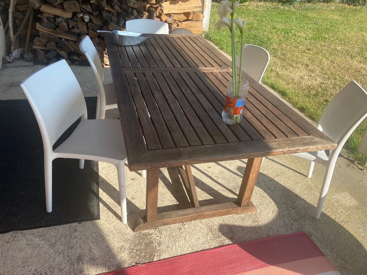 An outdoor dining area is depicted, featuring a wooden table with a slatted top. Four white chairs are arranged around the table, and a glass vase with fresh flowers is placed in the center. A grassy area is visible in the background.