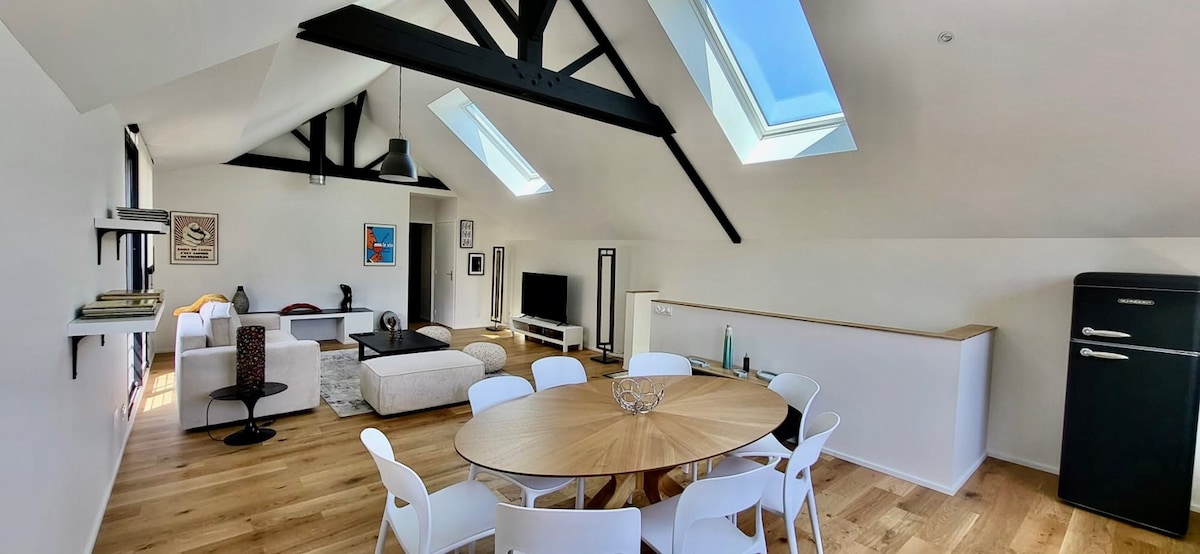 A spacious living area features high ceilings with exposed beams and skylights allowing natural light. A round wooden dining table is surrounded by white chairs. A comfortable seating arrangement is present, with a modern TV and shelving visible in the background.