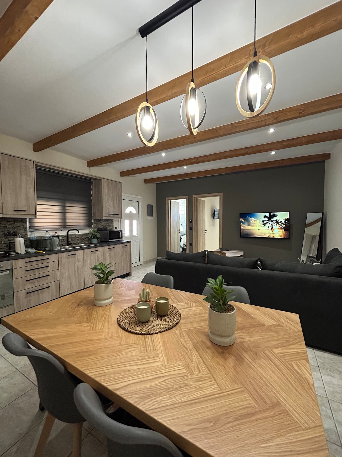 A modern kitchen and dining area features wooden beams overhead. A square table with a woven centerpiece and potted plants sits at the forefront. A cozy seating area with a dark sofa is visible, along with appliances and a flat-screen TV mounted on the wall.