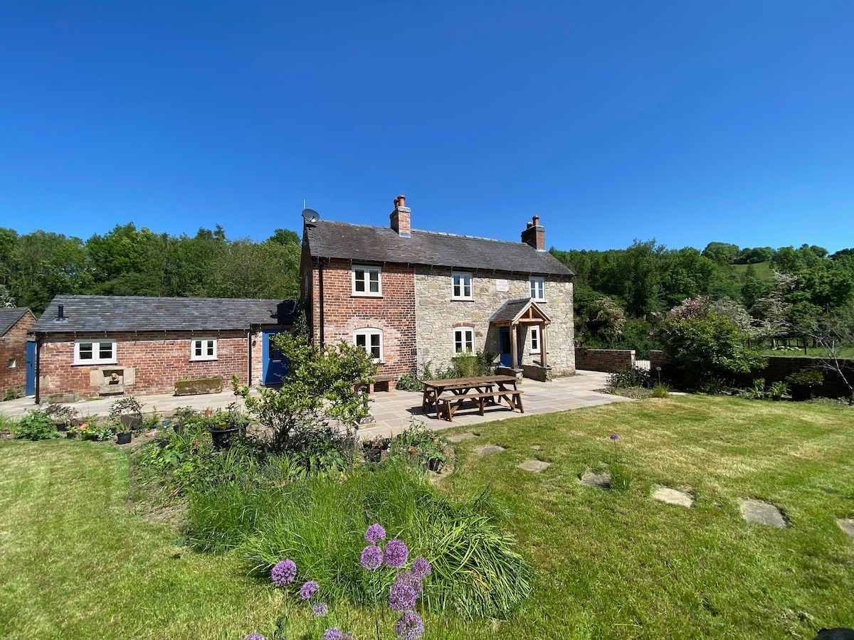 The charming cottage features a traditional brick facade and a welcoming entryway beneath an arch. The surrounding garden showcases lush greenery, colorful flowers, and a patio area with outdoor seating, all set against a backdrop of the serene countryside under a clear blue sky.