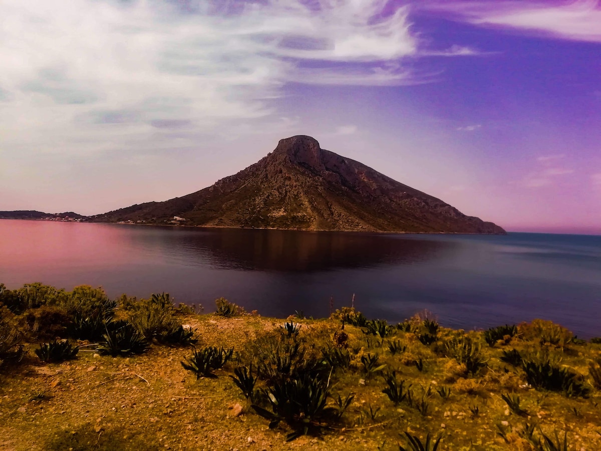 A serene coastal landscape presents a mountainous island rising from calm waters. The surrounding area features natural vegetation along the shore, with soft clouds drifting across the sky, creating a peaceful atmosphere.