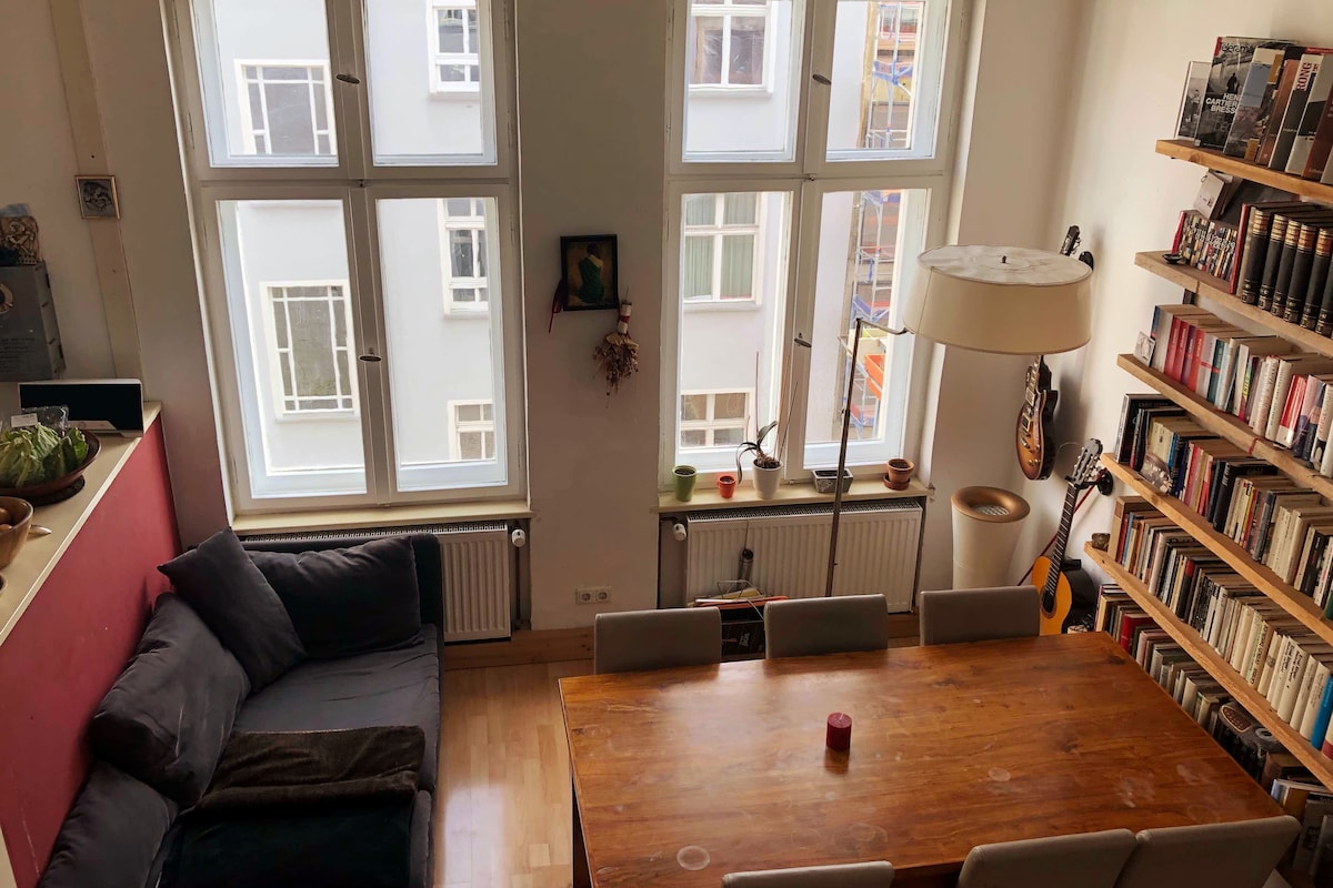 A spacious living and dining area is characterized by large windows allowing natural light. A long wooden dining table with seating for six is positioned in the center, while a cozy sofa is placed nearby. Shelves filled with books and a guitar are included in the décor.