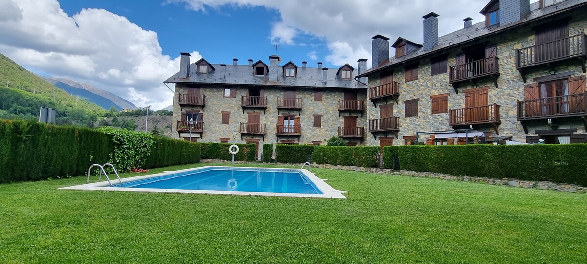 An outdoor area includes a well-maintained lawn and a blue swimming pool surrounded by greenery. The multi-story stone building features wooden balconies and multiple windows, providing a glimpse of the picturesque mountain landscape in the background.