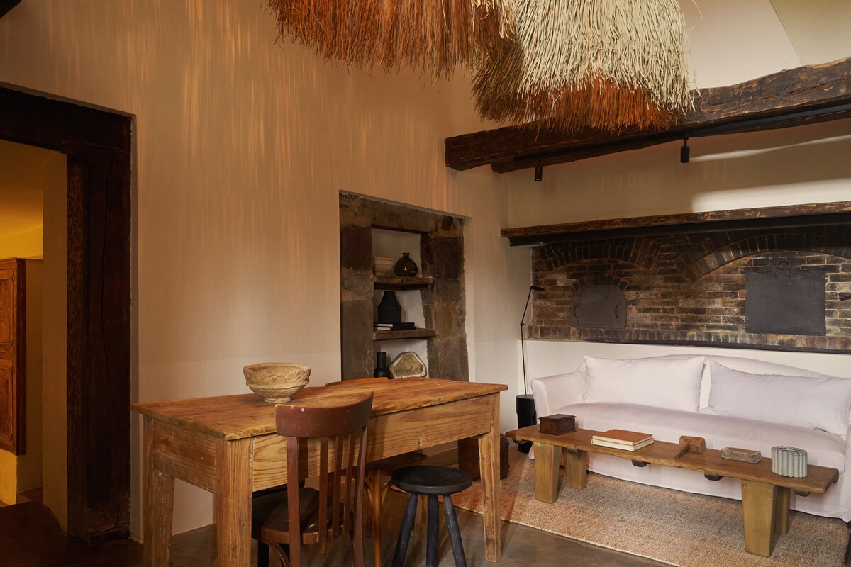 An inviting living area features a rustic wooden dining table surrounded by chairs. A cozy sofa rests against a brick wall, with decorative items displayed on a nearby shelf. The ceiling showcases an artistic straw hanging, adding texture to the space.