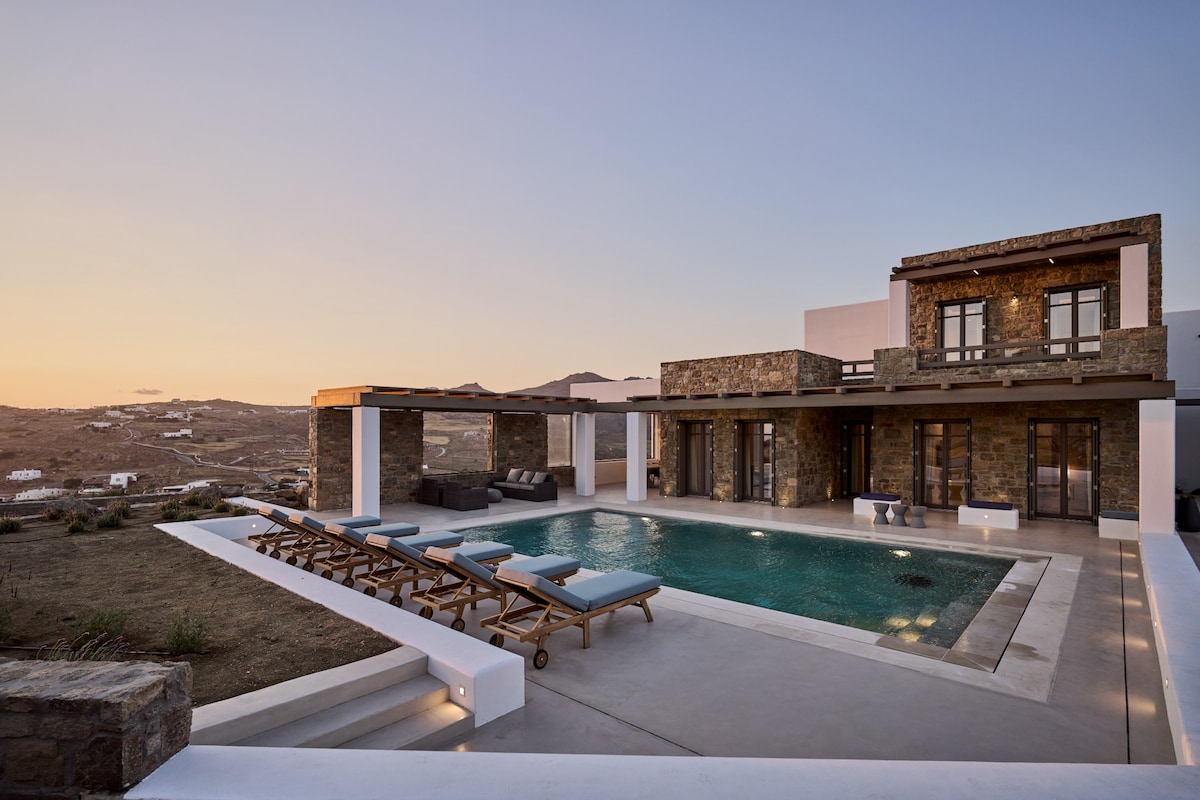 An inviting outdoor area features a rectangular swimming pool surrounded by wooden lounge chairs. The villa's stone facade is complemented by large windows and an expansive terrace, with a backdrop of gently rolling hills under a warm sunset sky.