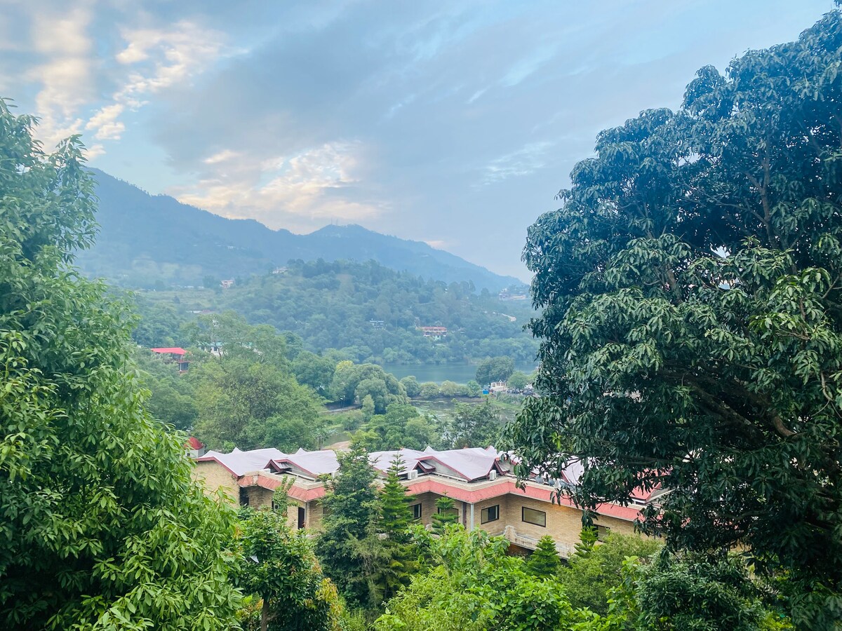 Lush greenery surrounds the area, with trees and hills visible in the distance. A view of nearby structures with red roofs blends seamlessly into the landscape, capturing a serene atmosphere. The sky features soft clouds, enhancing the tranquil backdrop.