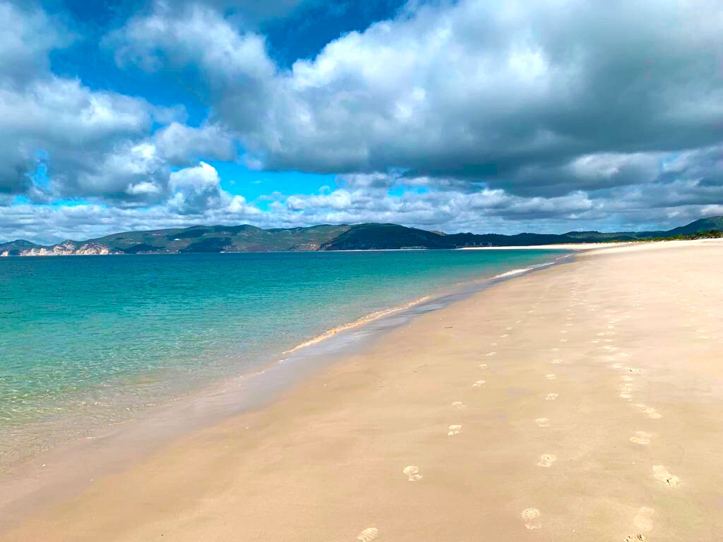 A tranquil beach scene is presented, featuring soft, golden sand leading to gently lapping waves. The water displays varying shades of blue, with distant green hills visible under a sky filled with soft, gray clouds and patches of blue. Footprints are scattered along the shoreline.