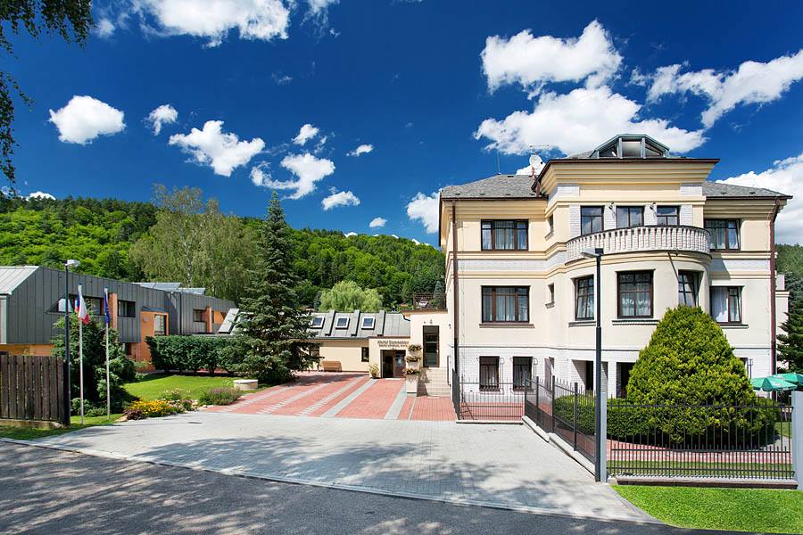 The exterior of the hotel is presented, surrounded by lush greenery and an open parking area. A multi-story building with a light-colored façade showcases a balcony and large windows. Sky features fluffy white clouds over a vibrant blue background, enhancing the property's inviting appearance.