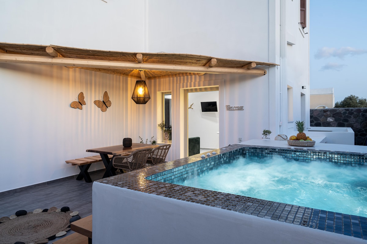 An inviting outdoor space features a tiled hot tub with bubbling water, surrounded by a wooden deck. A large dining table and chairs provide seating under a covered area, adorned with decorative wall elements. Soft lighting enhances the tranquil atmosphere of the villa.