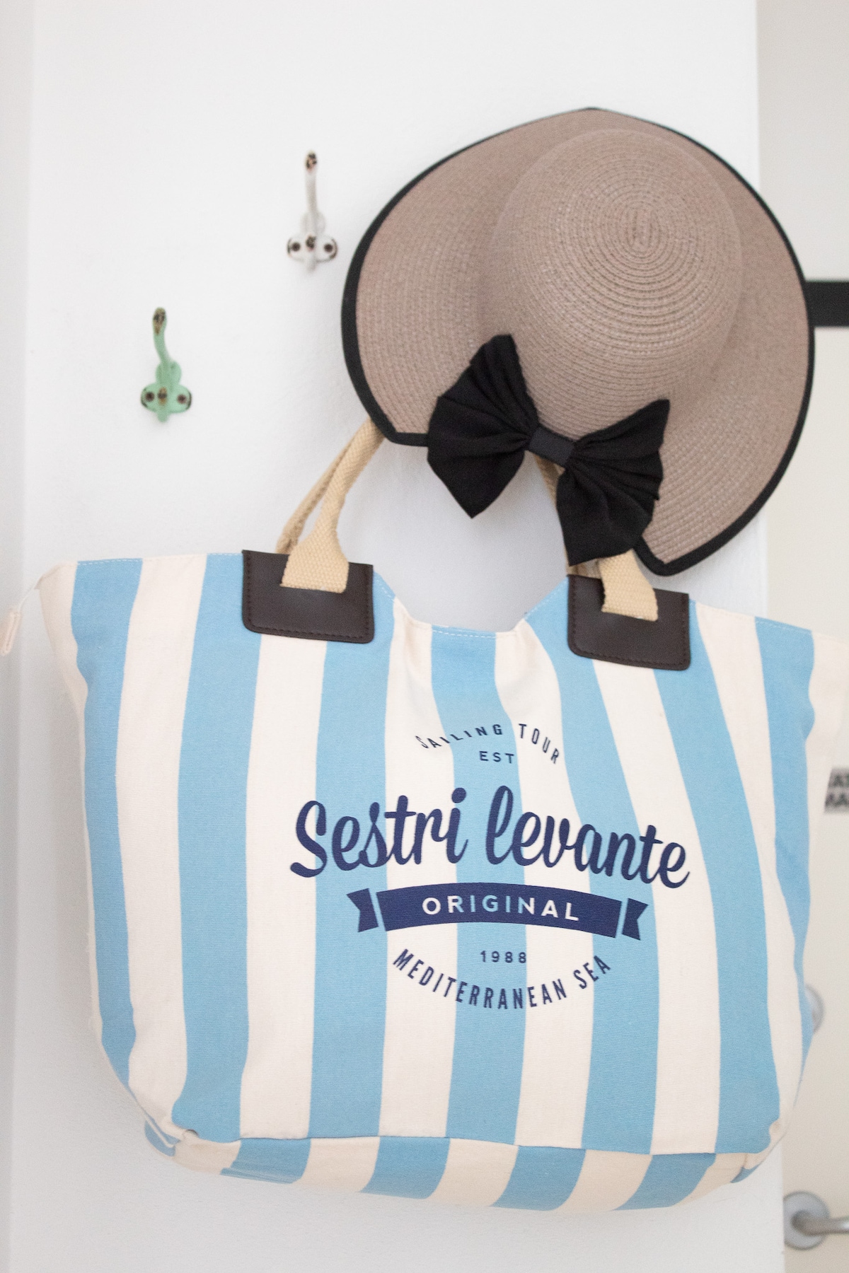 A striped beach bag in shades of blue and white is displayed against a light wall. A stylish sun hat, adorned with a black bow, hangs above the bag, evoking a coastal vibe. Decorative wall hooks are positioned to the side.