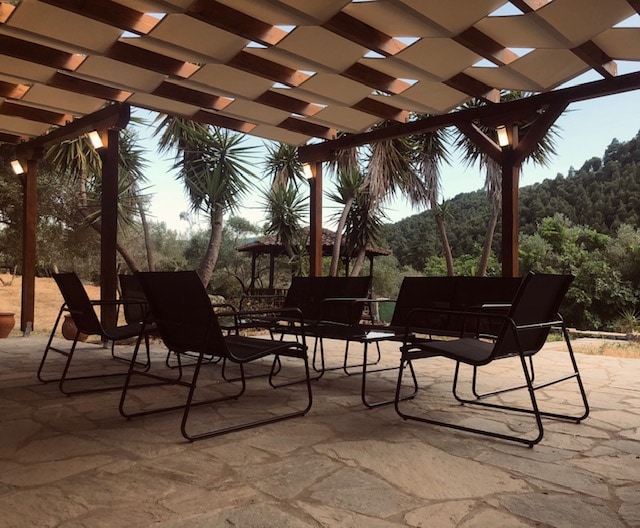 A spacious stone terrace is covered by a wooden pergola, offering shaded seating with several black lounge chairs arranged around. Lush greenery and palm trees are visible in the background, providing a serene atmosphere for relaxation.