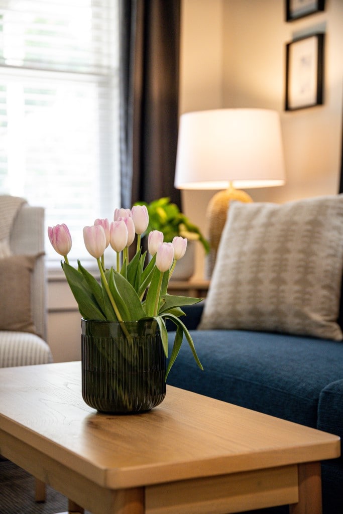 A decorative vase filled with pink tulips is placed on a wooden coffee table in a well-lit living area. Soft furnishings, including a light-colored accent chair and a dark blue couch, are visible, complemented by a warm lamp and curtains filtering natural light.