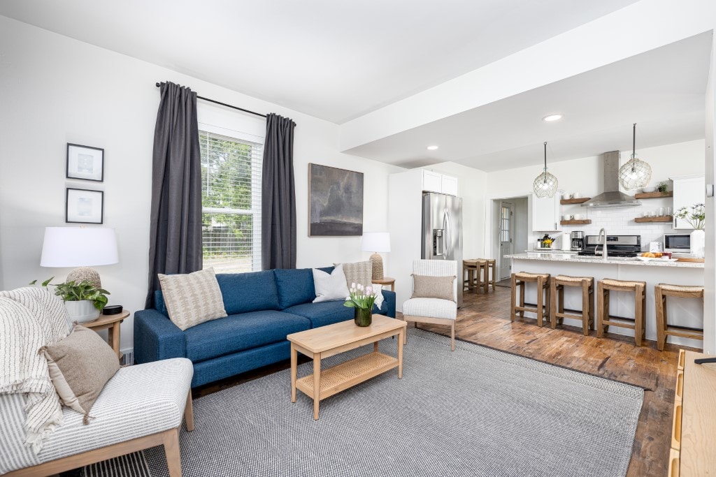 A spacious living area features a blue sofa and a neutral armchair, complemented by a wooden coffee table. Natural light enters through a window with sheer curtains. An open kitchen with a large island and modern appliances is visible in the background, creating a cohesive living space.