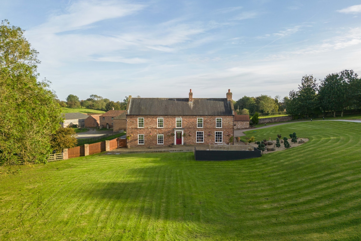 A spacious country estate is showcased, featuring a classic brick facade with multiple windows. Surrounding lawns are neatly trimmed, complemented by manicured gardens. Rolling hills and trees are visible in the background, offering a serene rural setting.