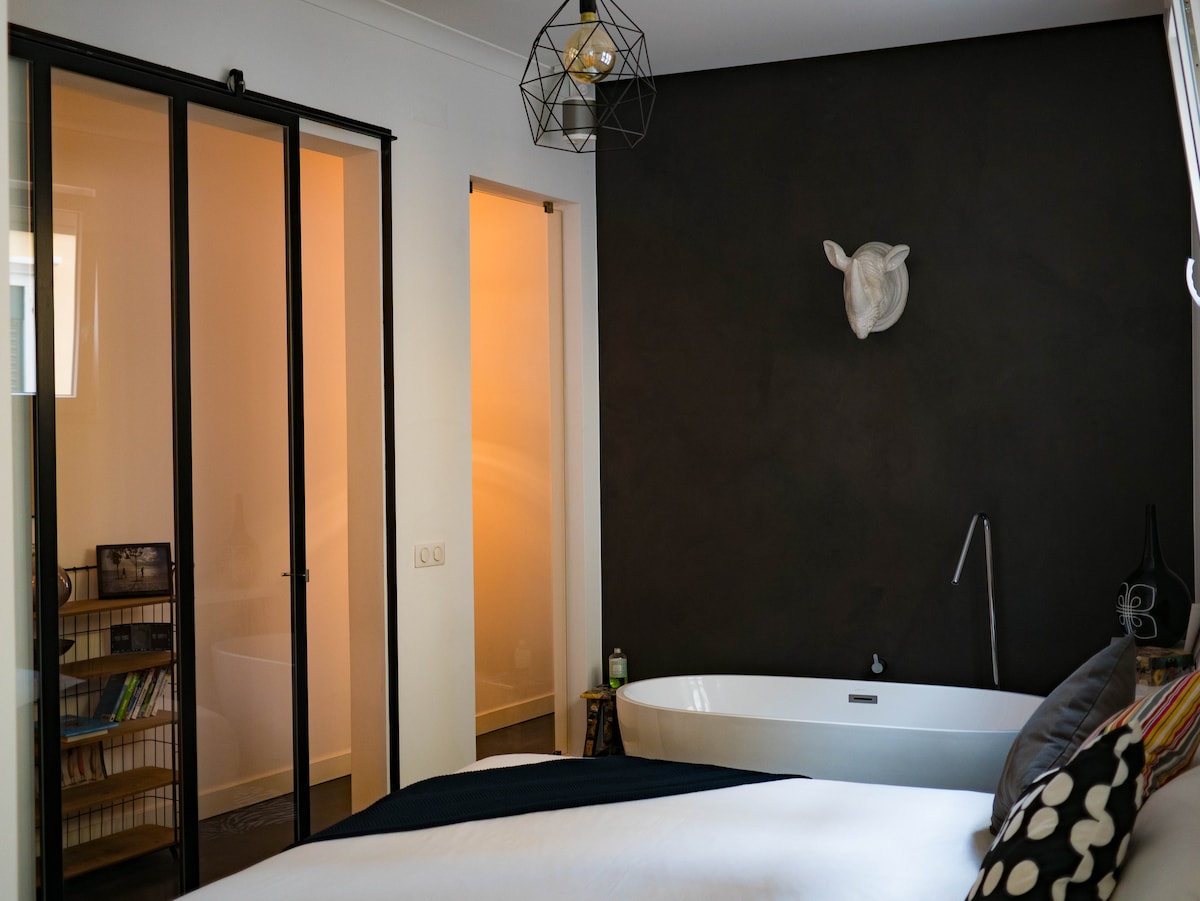 A stylish bedroom features a spacious bed with a crisp white duvet, complemented by decorative pillows. A modern freestanding bathtub is positioned next to the wall, enhancing the design. Nearby, glass sliding doors reveal an adjoining room, while soft lighting emanates from a geometric pendant lamp.